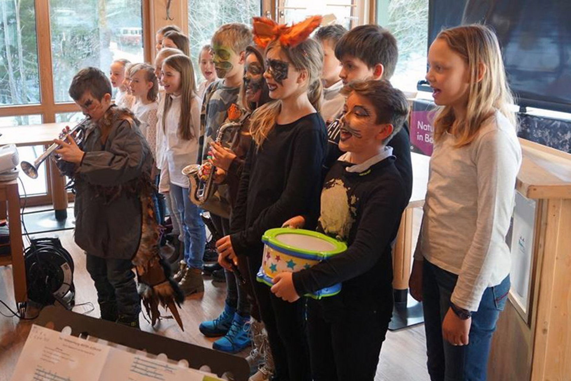 Schülerinnen und Schüler, teilweise mit geschminkten Gesichtern, singen und musizieren in einem Raum.