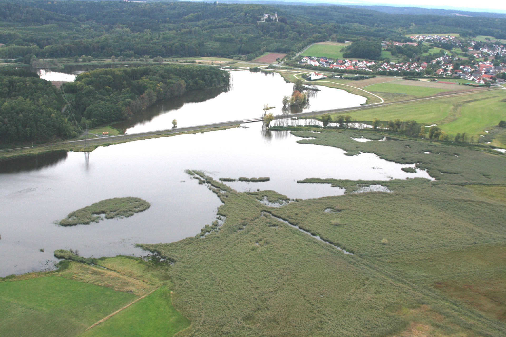 Luftbild einer Landschaft mit überschwemmten Wiesen. Im Hintergrund eine Ortschaft und bewaldete Hänge.